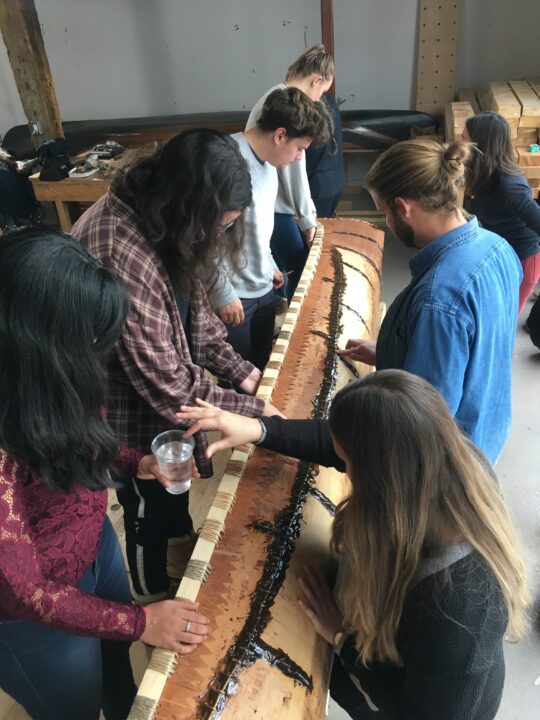 Photograph of students pouring water on the side of a birch bark canoe