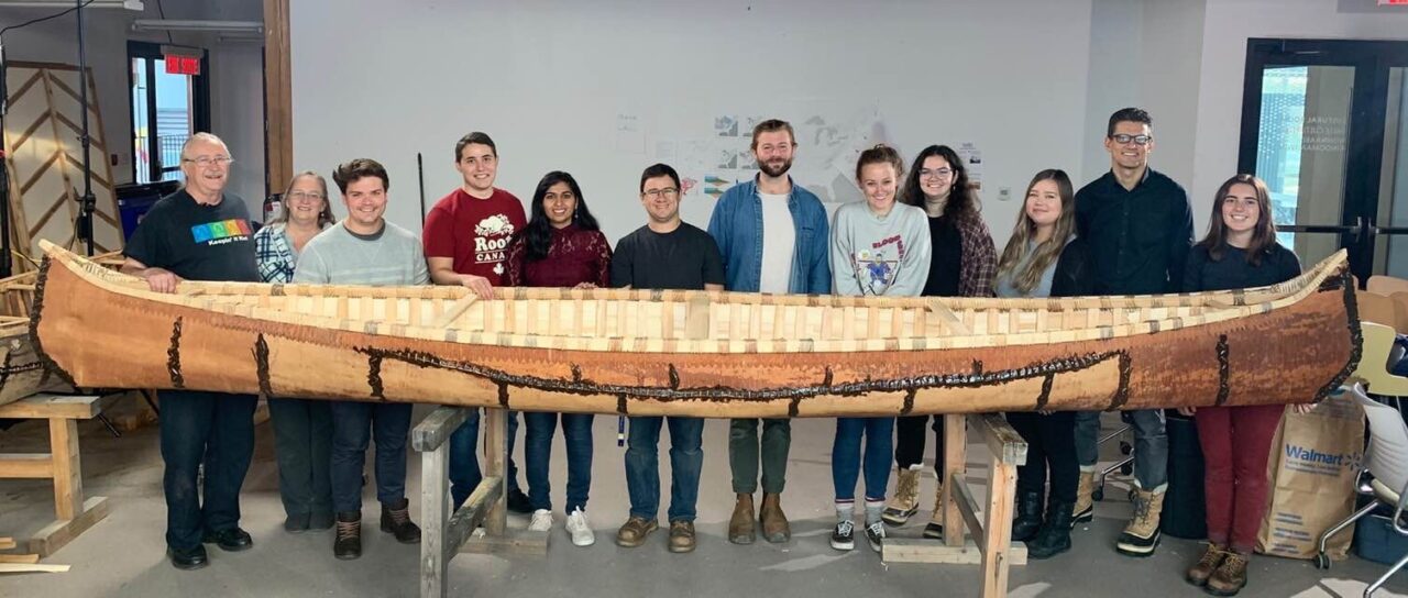 Photograph of students and staff standing behind the birch bark canoe they built