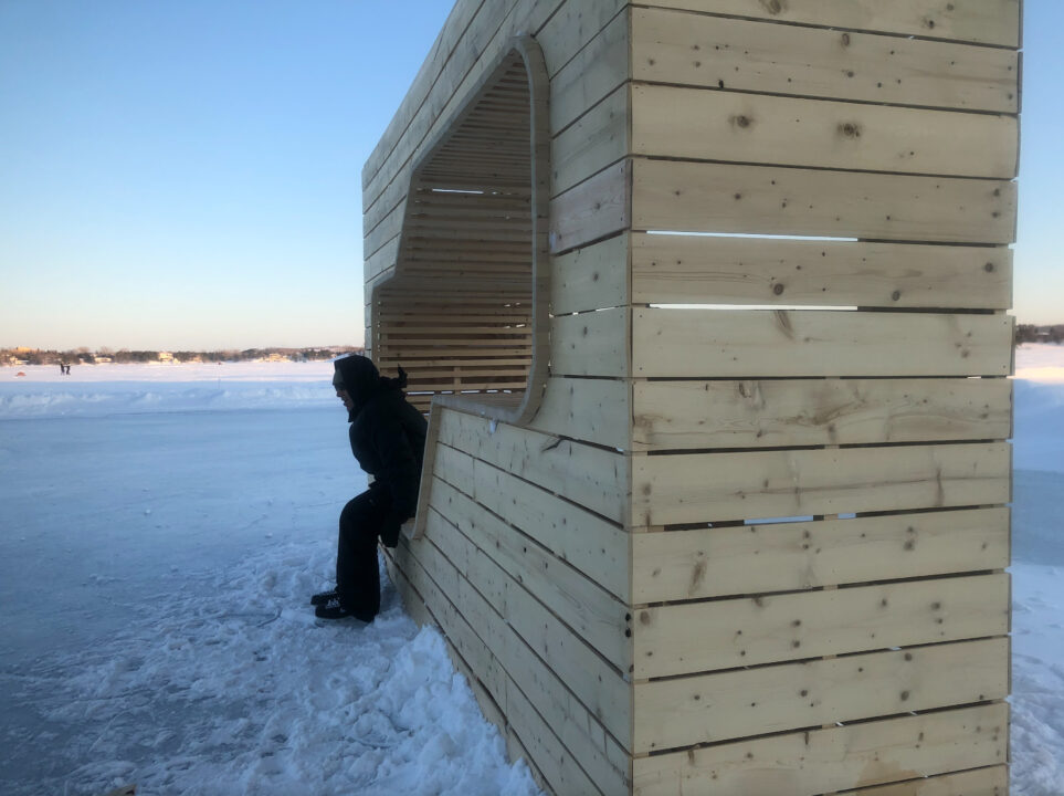 Photograph of a person sitting in a wooden ice warming station on a skating path in winter